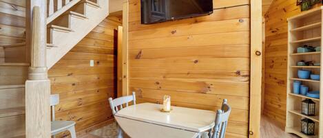 Un intérieur en bois avec un escalier, une table entourée de chaises, un téléviseur fixé au mur et une étagère avec des bols bleus.