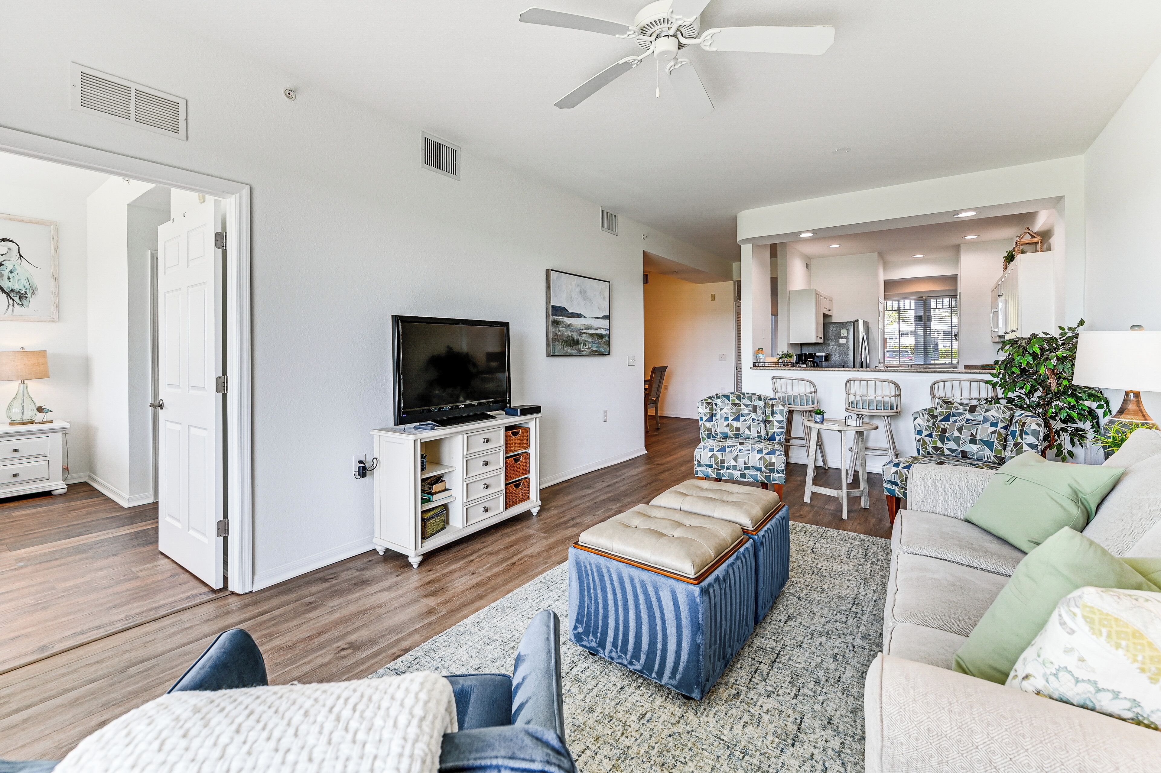 A spacious living room with a television, ottoman, and sofa, a kitchen in the background, and a ceiling fan.