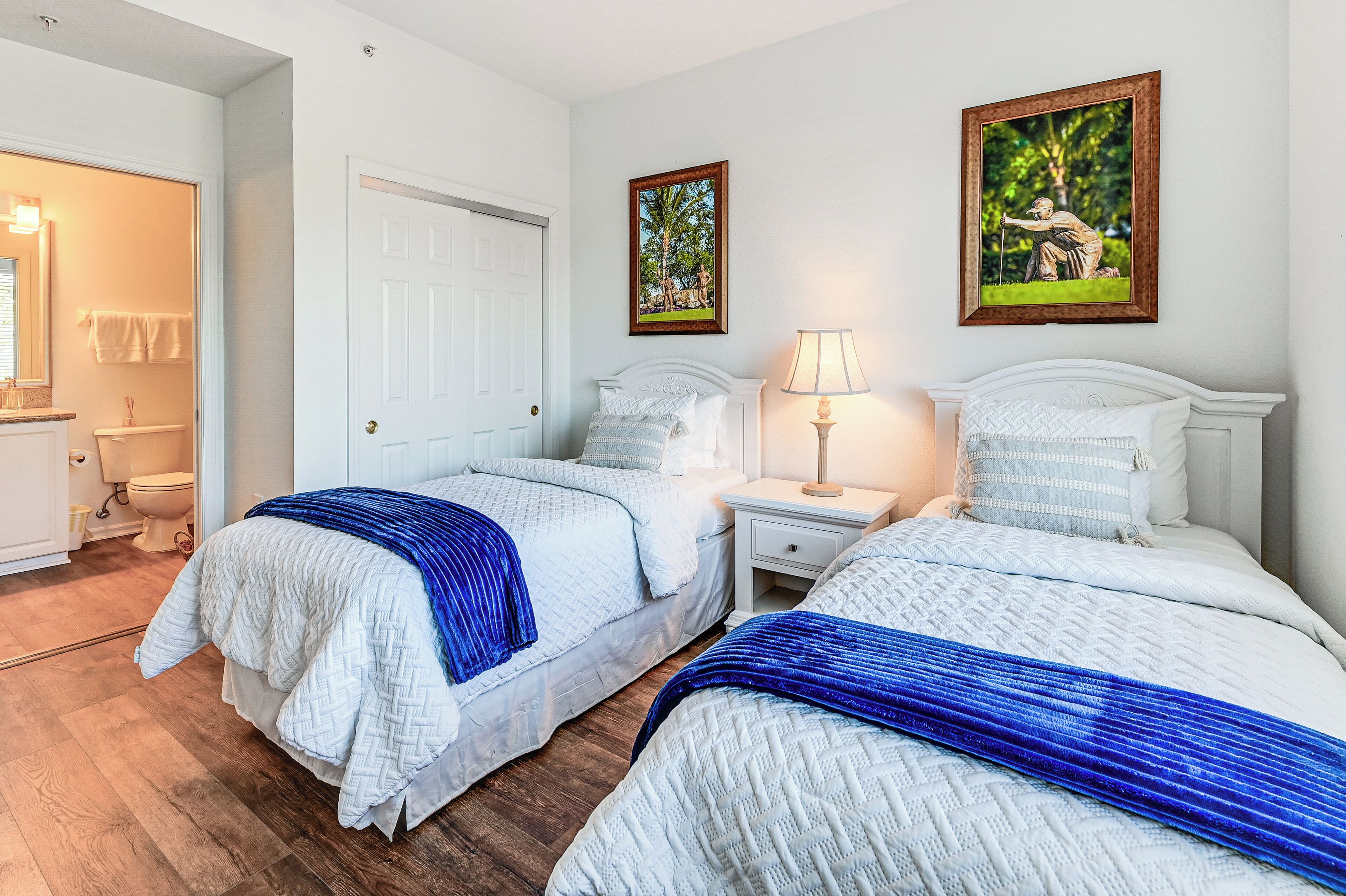 A hotel room with two beds, a white door, a nightstand with a lamp, and a framed picture on the wall.