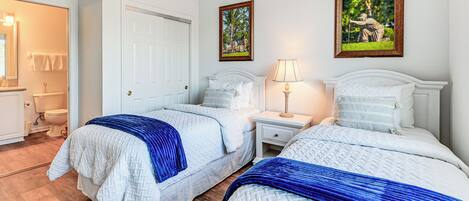 A hotel room with two beds, a white door, a nightstand with a lamp, and a framed picture on the wall.