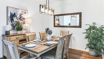 A dining area with a wooden table set for six, surrounded by chairs. There is a large abstract painting on the wall, a mirror, and a potted plant.