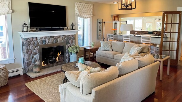 A living room with a stone fireplace, a flat-screen TV, a sofa set, and a kitchen area visible through an open door. - 5BR Lake-view Home w/ Pool & Seasonal Hot Tub (Beulah)