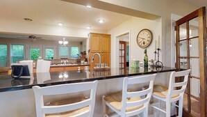 A modern kitchen with a central island, white bar stools, wooden cabinets, and a large clock on the wall. - 5BR Lake-view Home w/ Pool & Seasonal Hot Tub (Beulah)