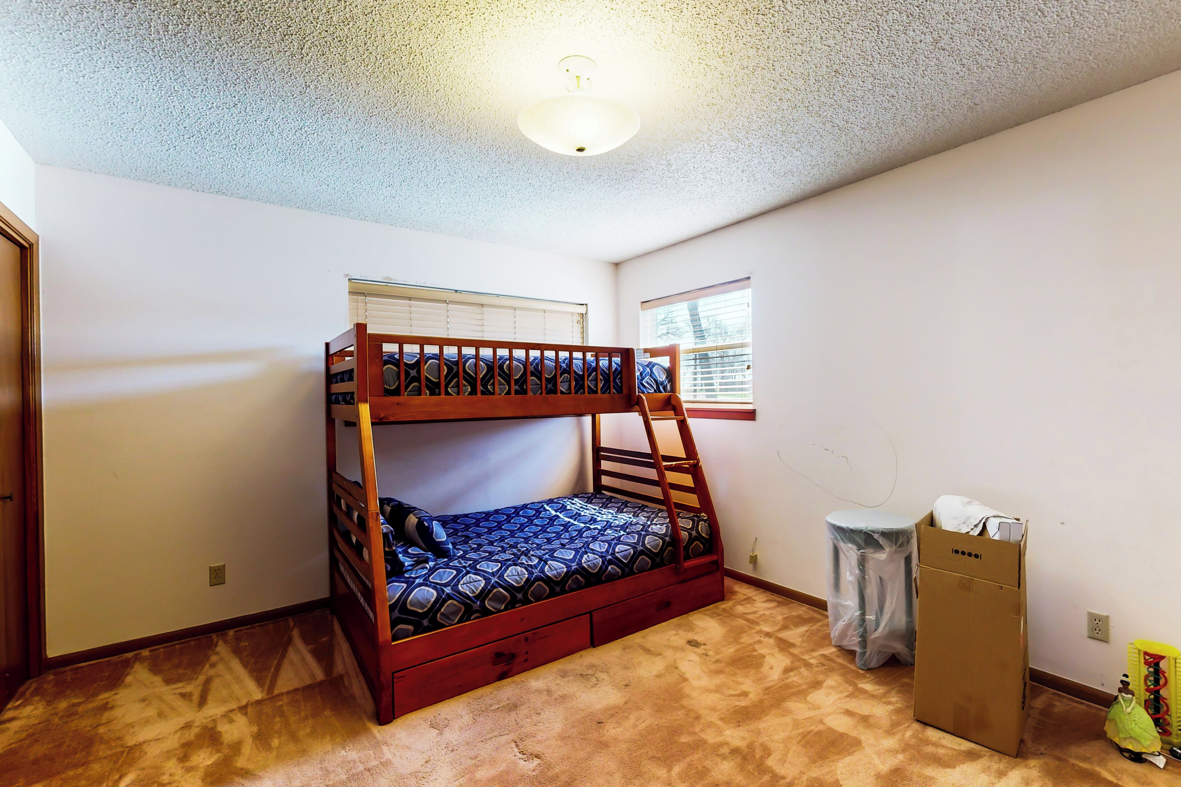 Una cama litera en una habitación con piso alfombrado, una ventana con persianas y una lámpara en el techo.
