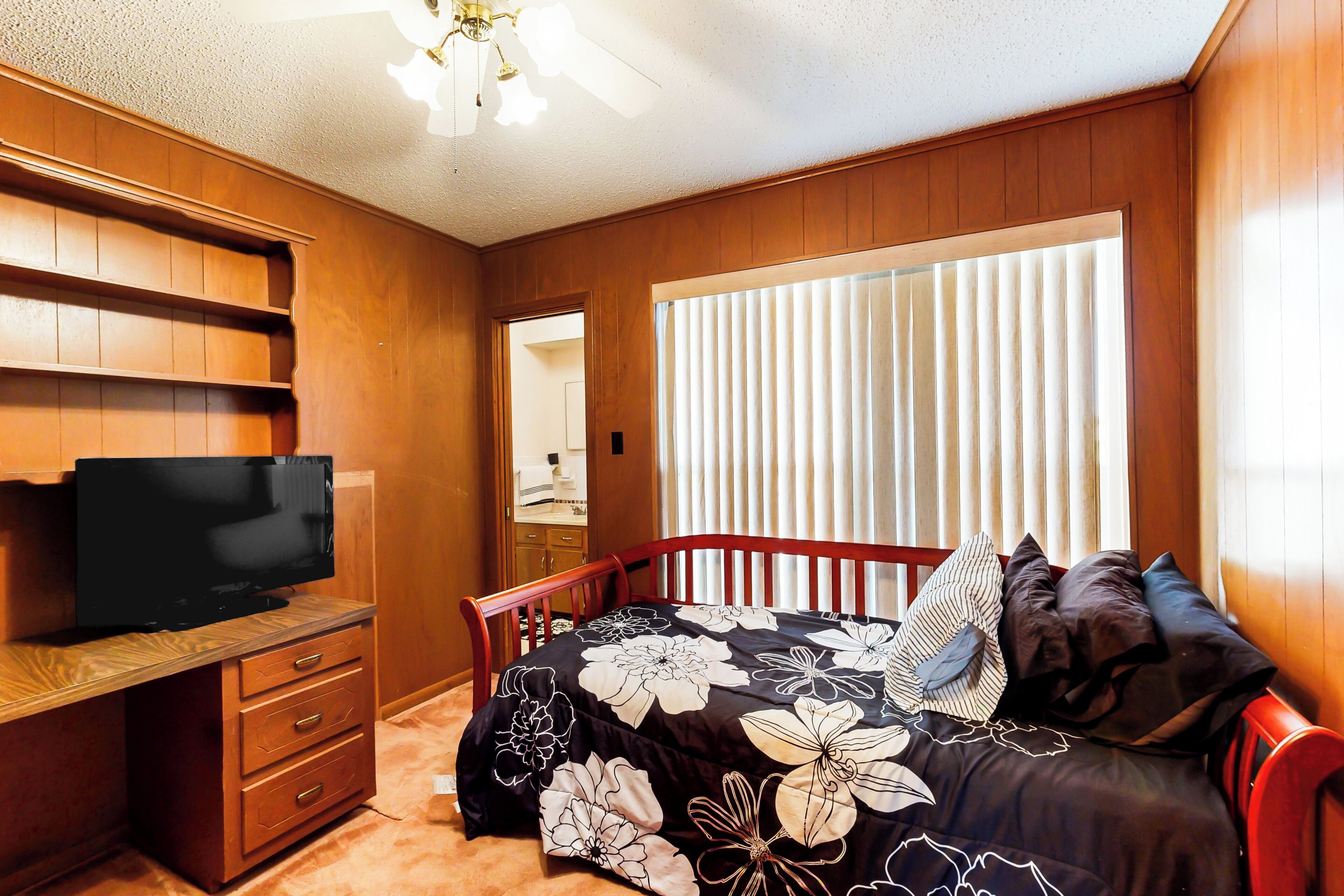 A bedroom with a wooden headboard, a bed with floral bedding, a television on a wooden dresser, and a window with blinds.