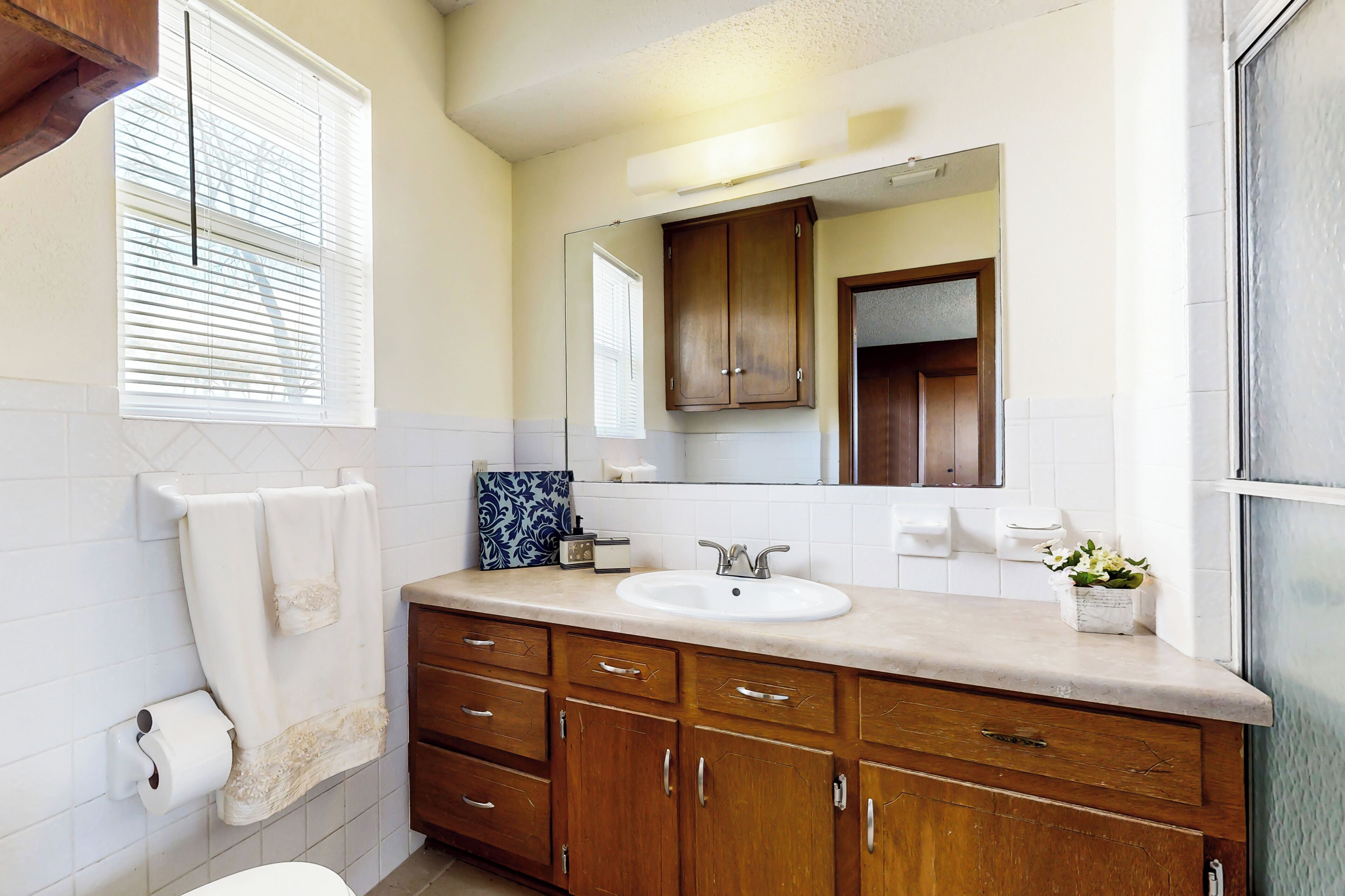 Une salle de bain avec un lavabo blanc, des meubles en bois, un miroir et une fenêtre avec des stores.
