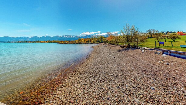 A pebbly lakeshore with a bench and a sign, surrounded by trees and mountains in the background. - Soft Sunsets & Still Water at Flathead Condo (Polson)