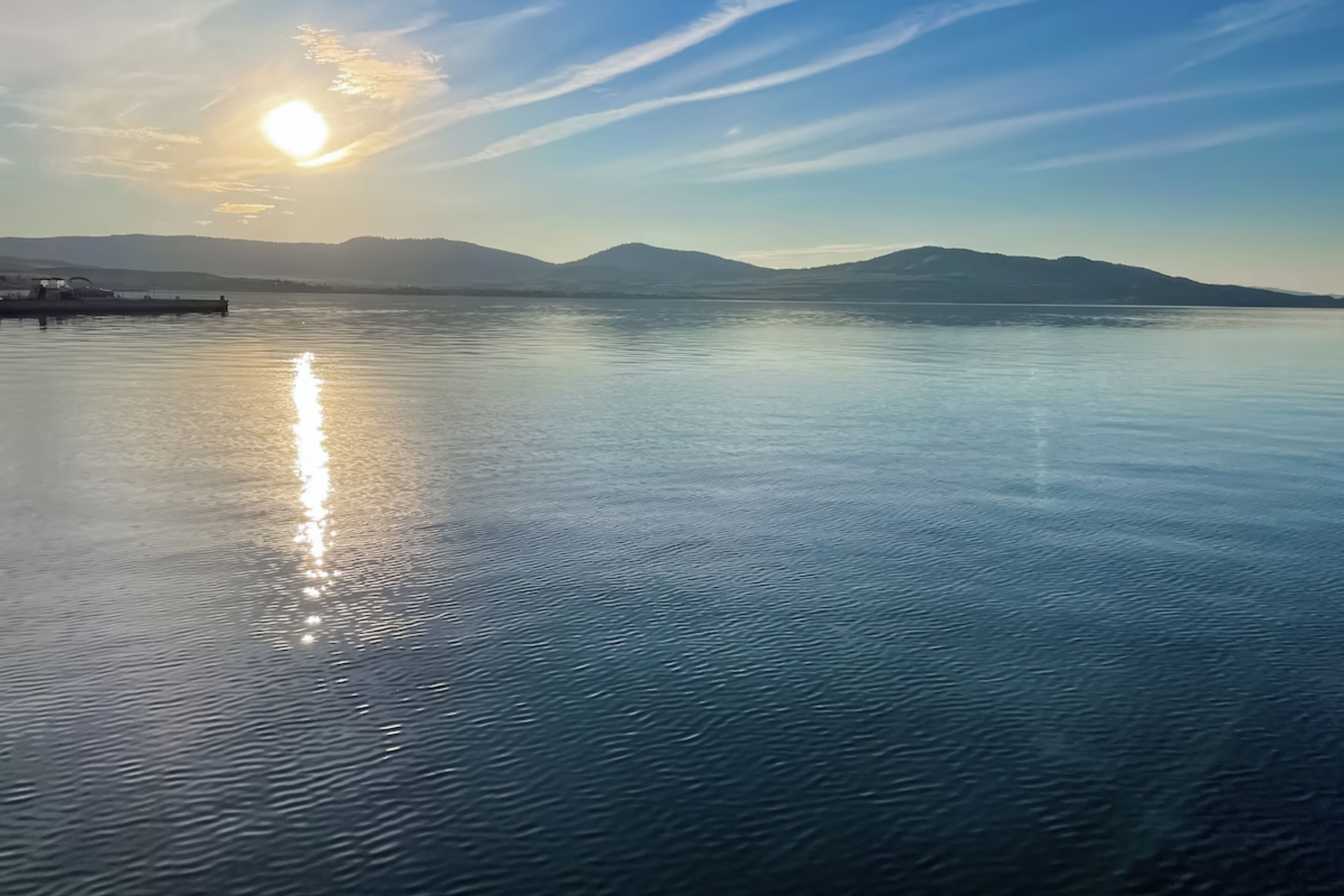 Une étendue d’eau calme où se reflète le soleil, avec des montagnes en arrière-plan et un quai sur la gauche.