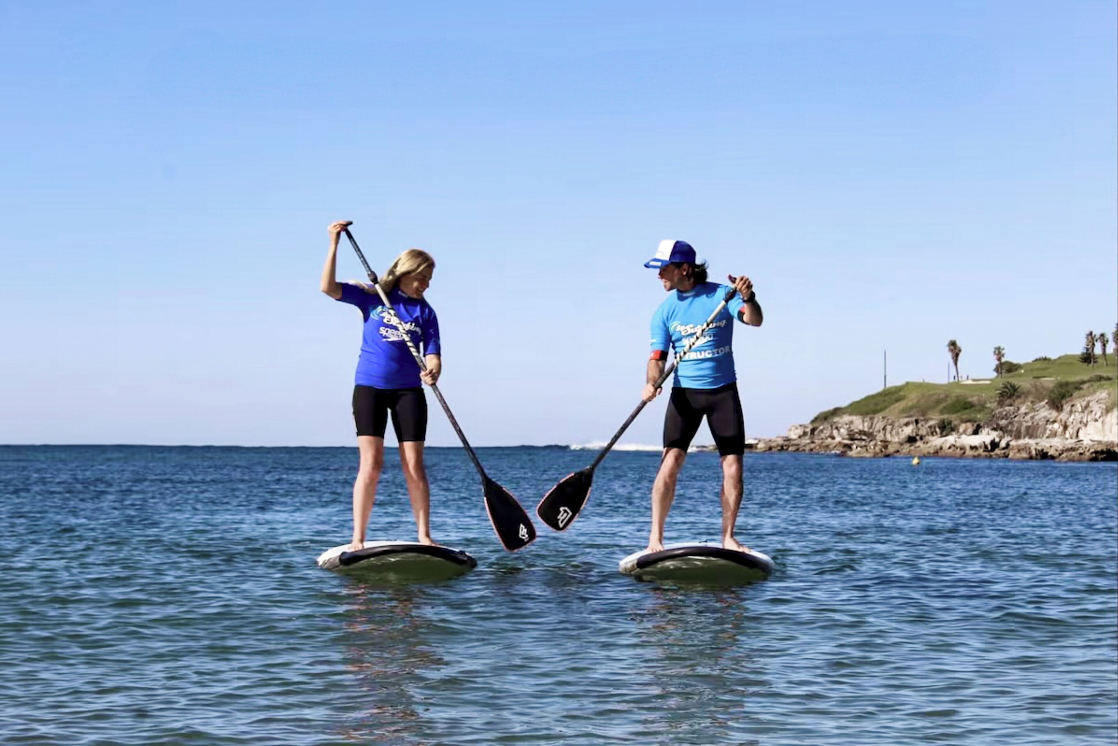 Two people stand-up paddleboarding on a calm sea with a clear sky.