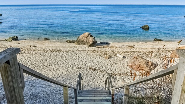 Una escalera de madera que baja a una playa de arena con agua azul cristalina y rocas.