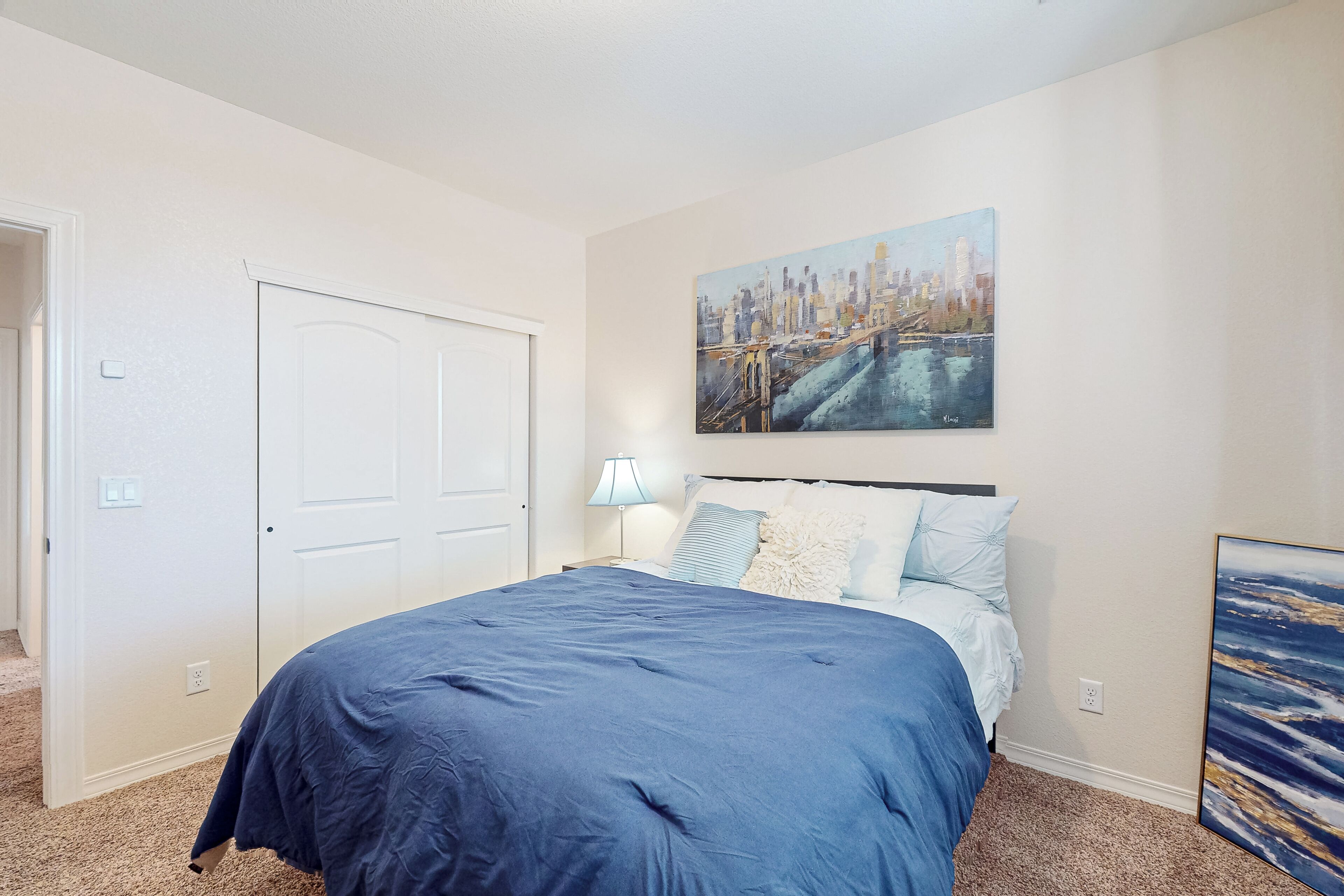 A bedroom with a large bed, a cityscape painting above it, and a door to the left.