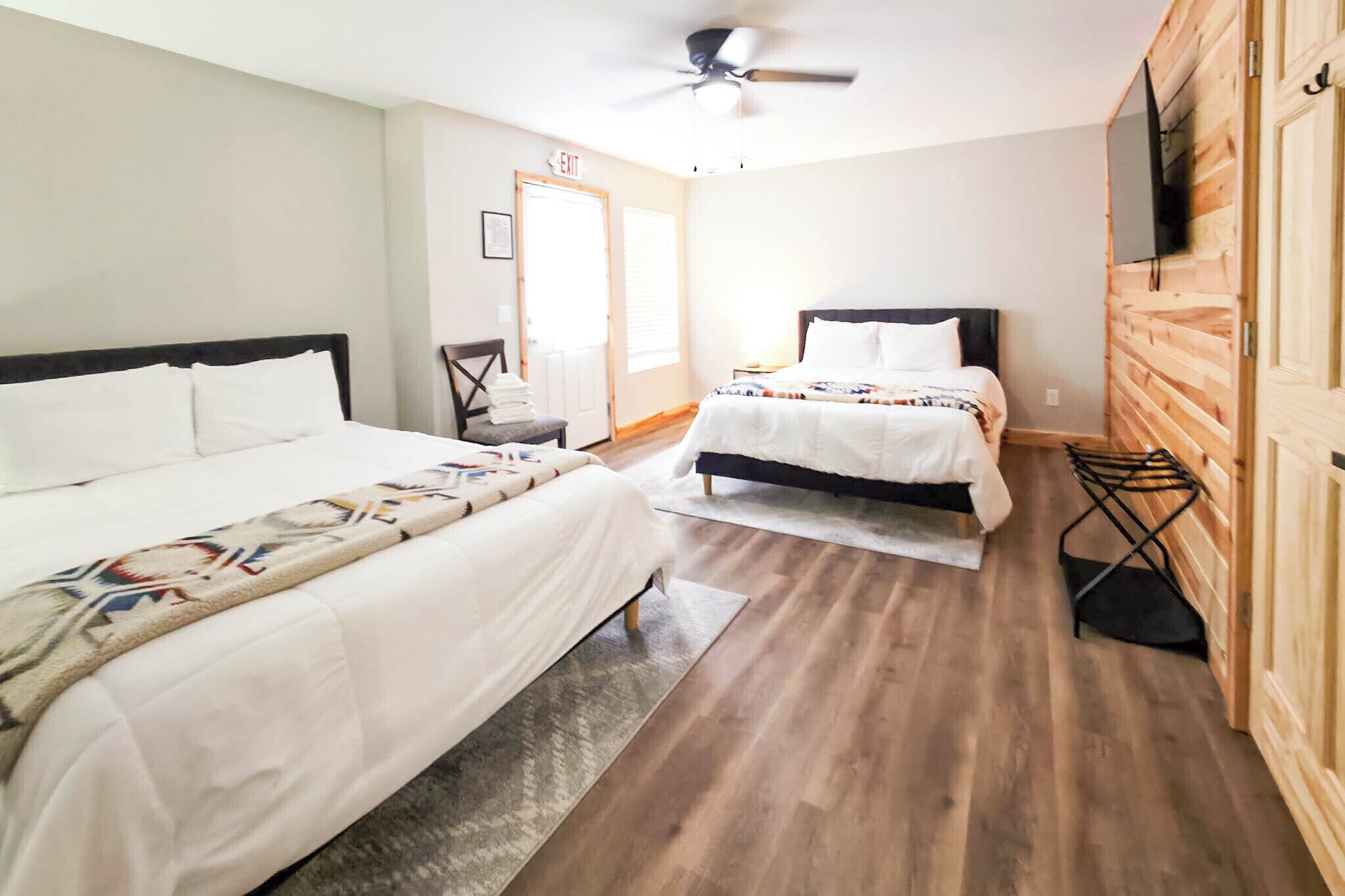 A hotel room with two beds, a ceiling fan, a TV mounted on the wall, and a wooden floor.