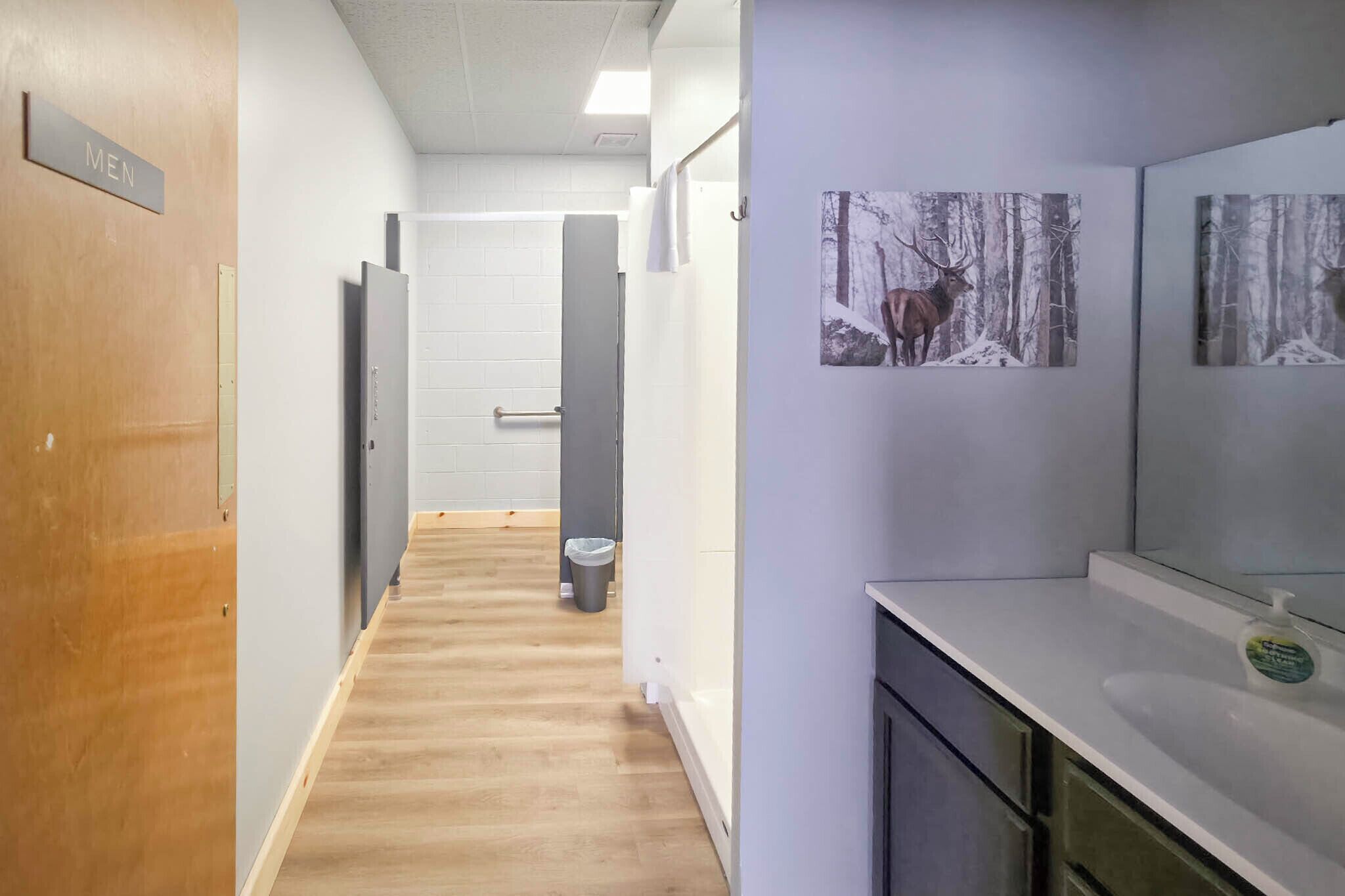 A hallway with a wooden floor, a men's restroom sign, a trash bin, and a sink with a soap dispenser.
