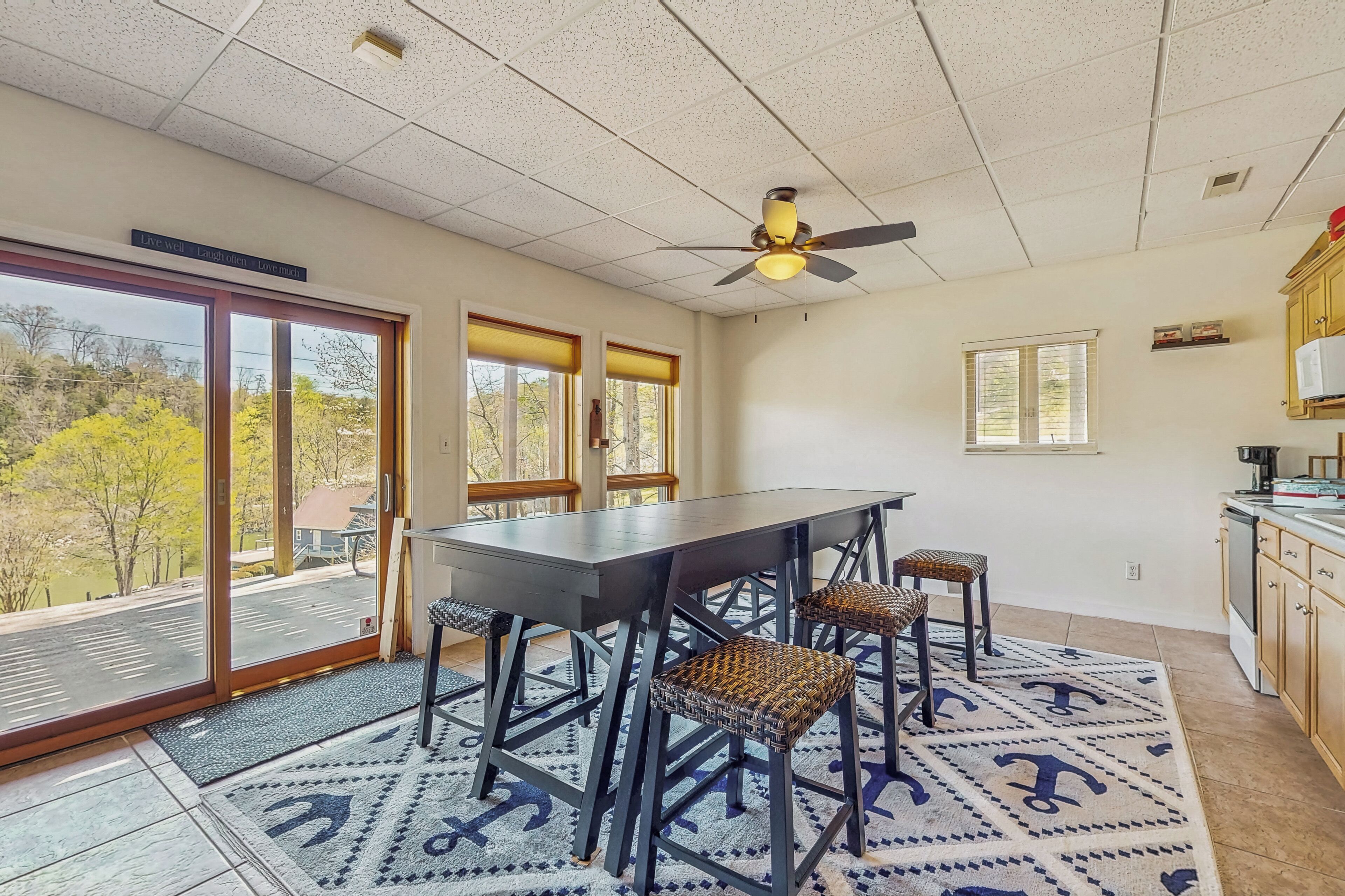 A dining area with a table and chairs, a ceiling fan, and a view of trees through large windows.