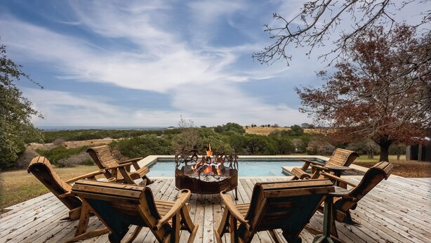 A wooden deck with outdoor furniture and a fire pit, overlooking a pool and scenic landscape. - 5BR Luxury Home w/ Pool & Wrap-around Porch (Glen Rose)