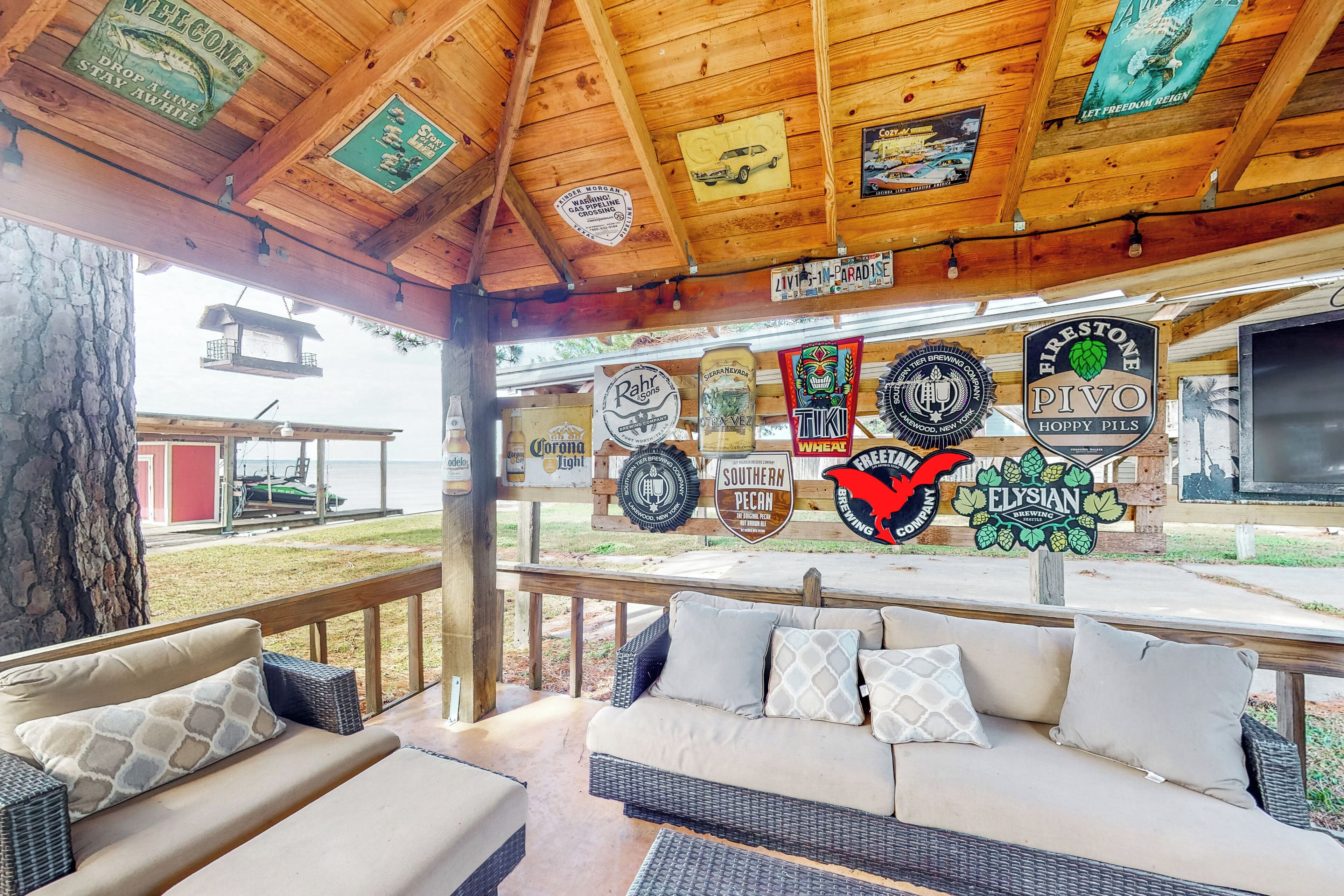 A covered outdoor seating area with wicker furniture and a wooden ceiling displaying various brewery logos.