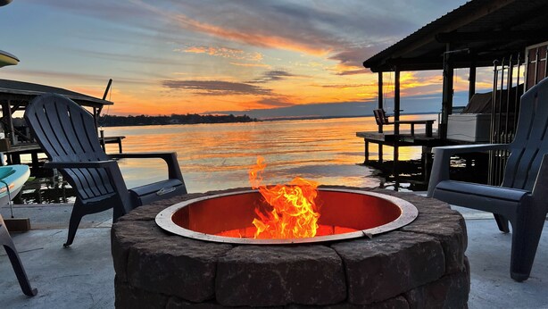 A stone fire pit with a view of a sunset over a body of water, surrounded by Adirondack chairs and a covered seating area. - 'harbor Haven' w/ Dock: Lake Livingston Home (Livingston)