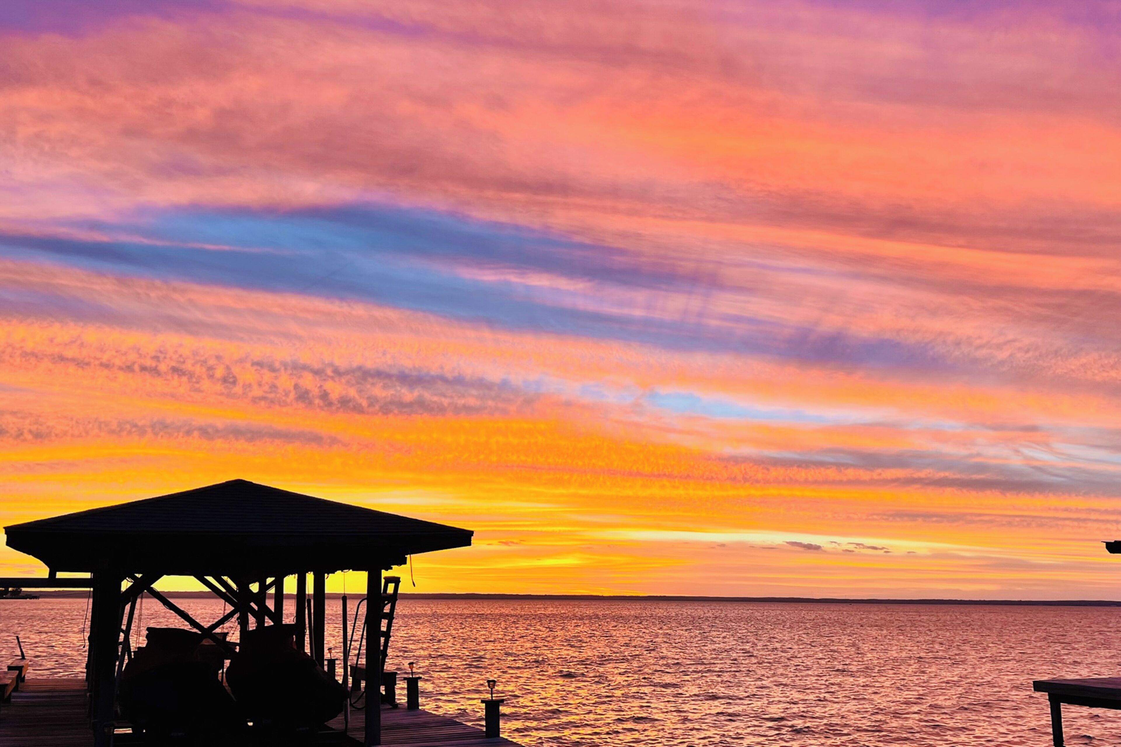 A pier with a covered seating area overlooking a sunset over the water.