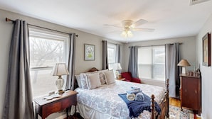Ein Schlafzimmer mit einem Bett, Nachttischen, einem Schreibtisch, einem Stuhl, einem Deckenventilator und einem Fenster mit Vorhängen.