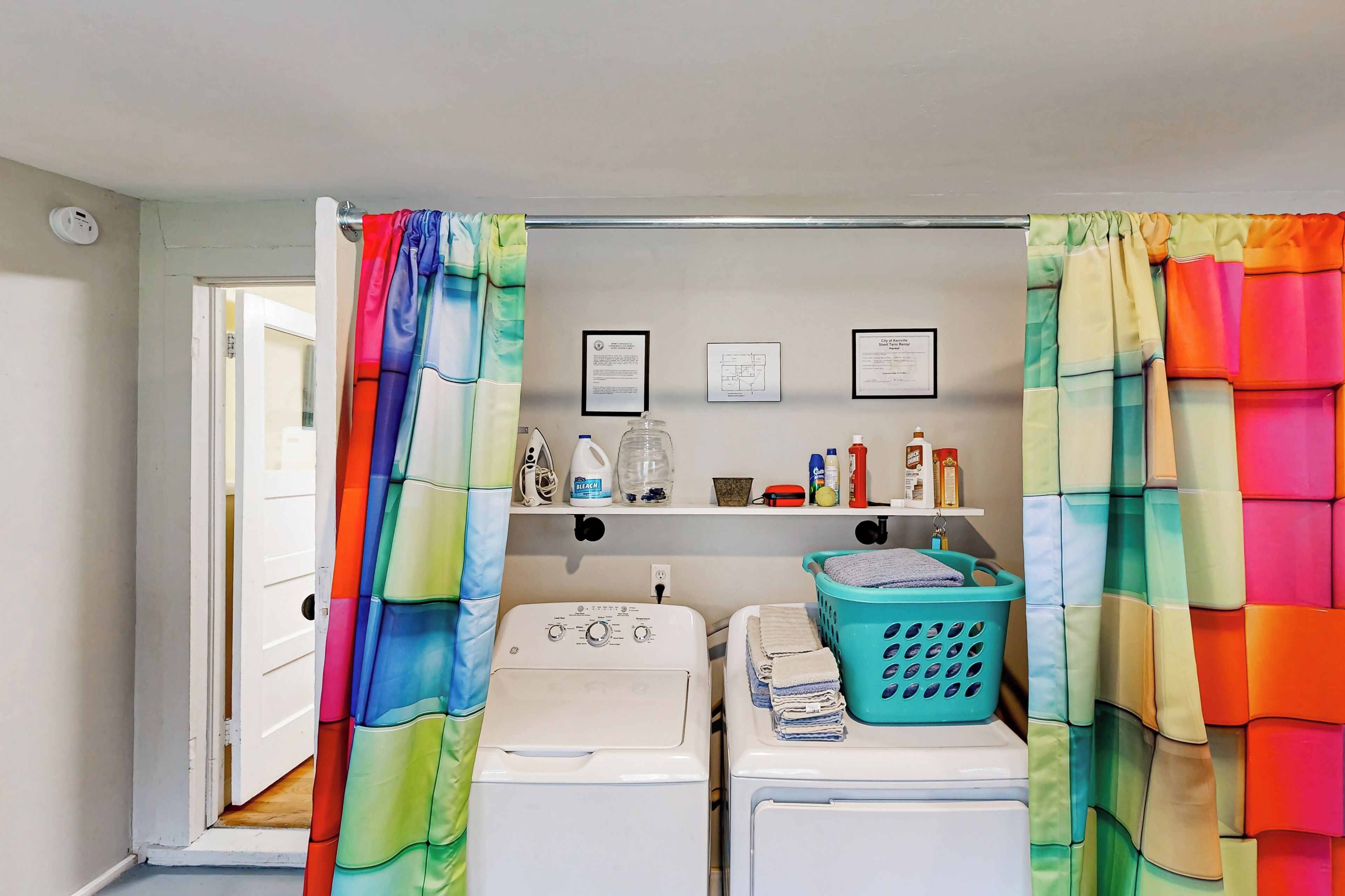 Un lavadero con dos lavadoras, un estante con productos de limpieza y una puerta que da a otra habitación.