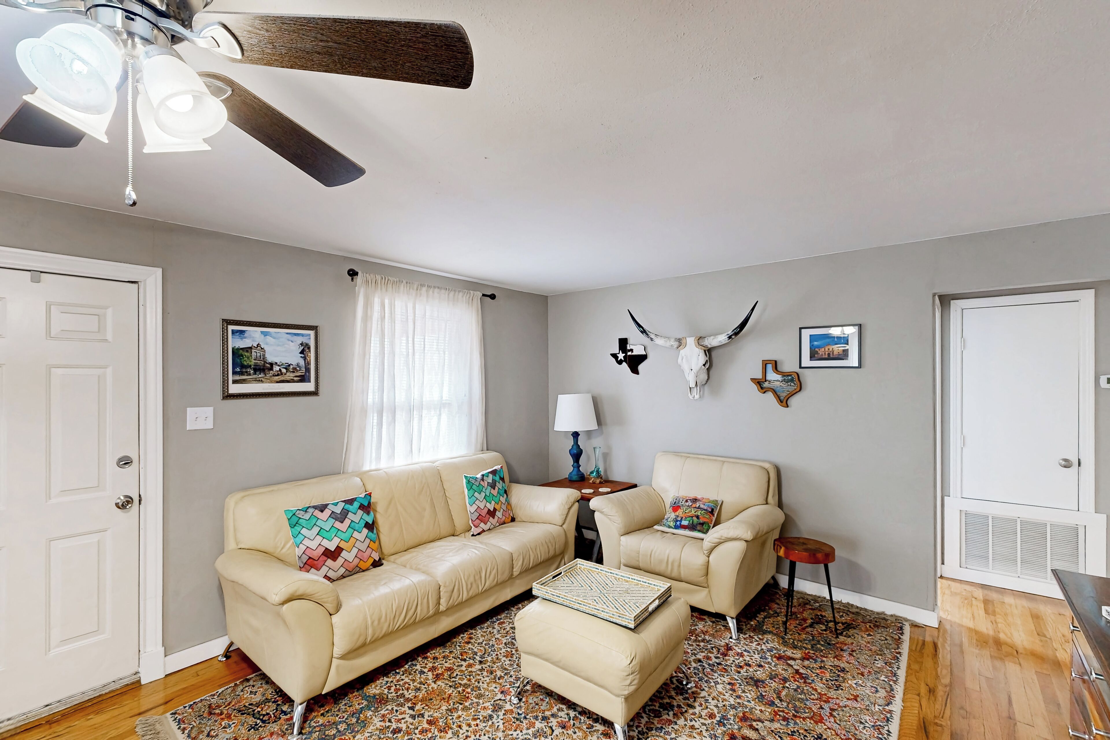 A living room with a beige leather sofa, a matching ottoman, a patterned rug, a ceiling fan, and a wall-mounted deer head decoration.