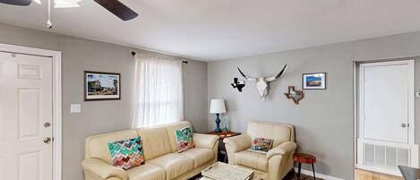 A living room with a beige leather sofa, a matching ottoman, a patterned rug, a ceiling fan, and a wall-mounted deer head decoration.