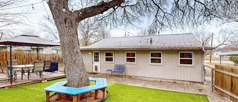 Un patio trasero con un árbol, un banco azul, un porche cubierto y una casa con techo de metal.