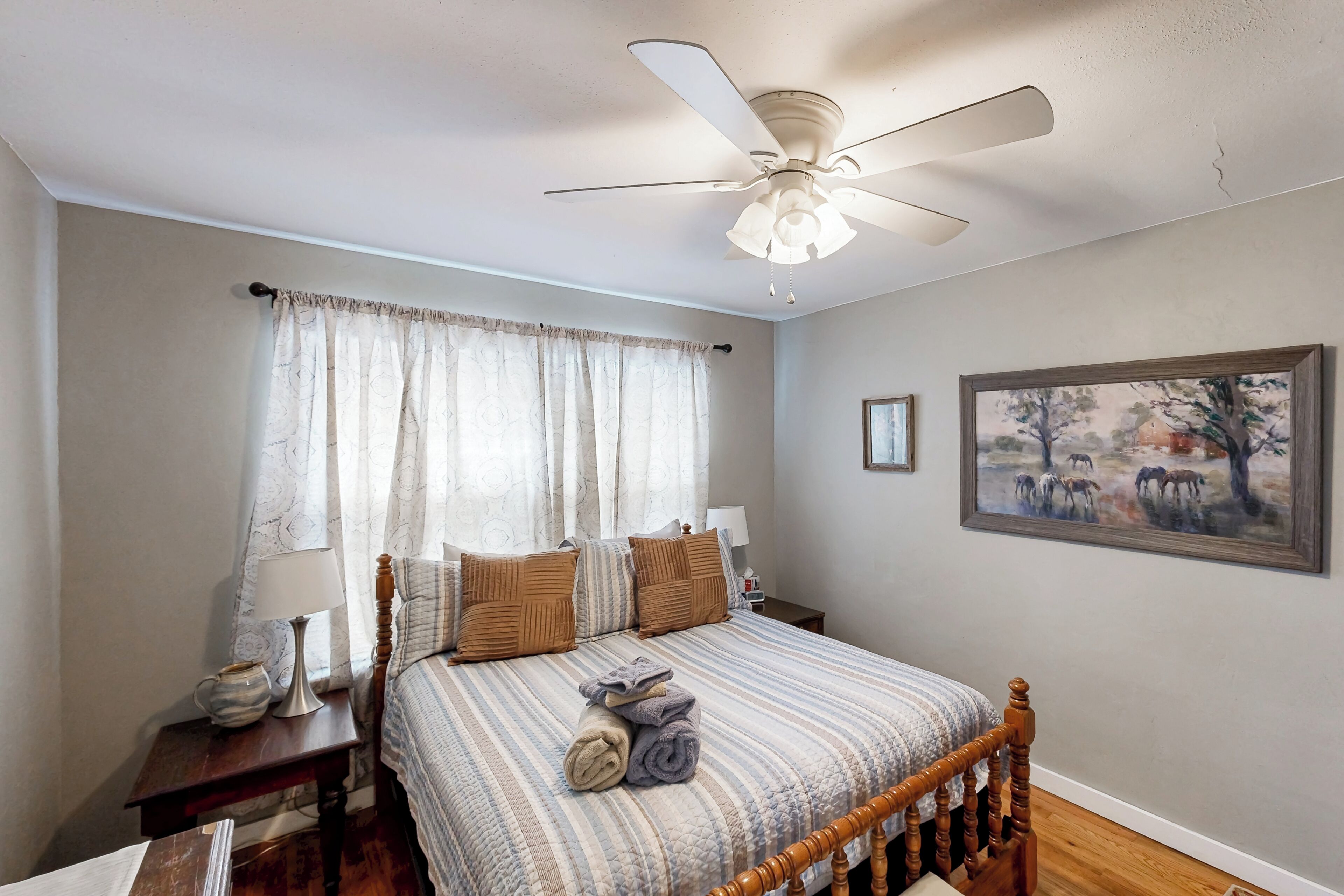 A bedroom with a bed, a nightstand, a lamp, a ceiling fan, and a painting on the wall.