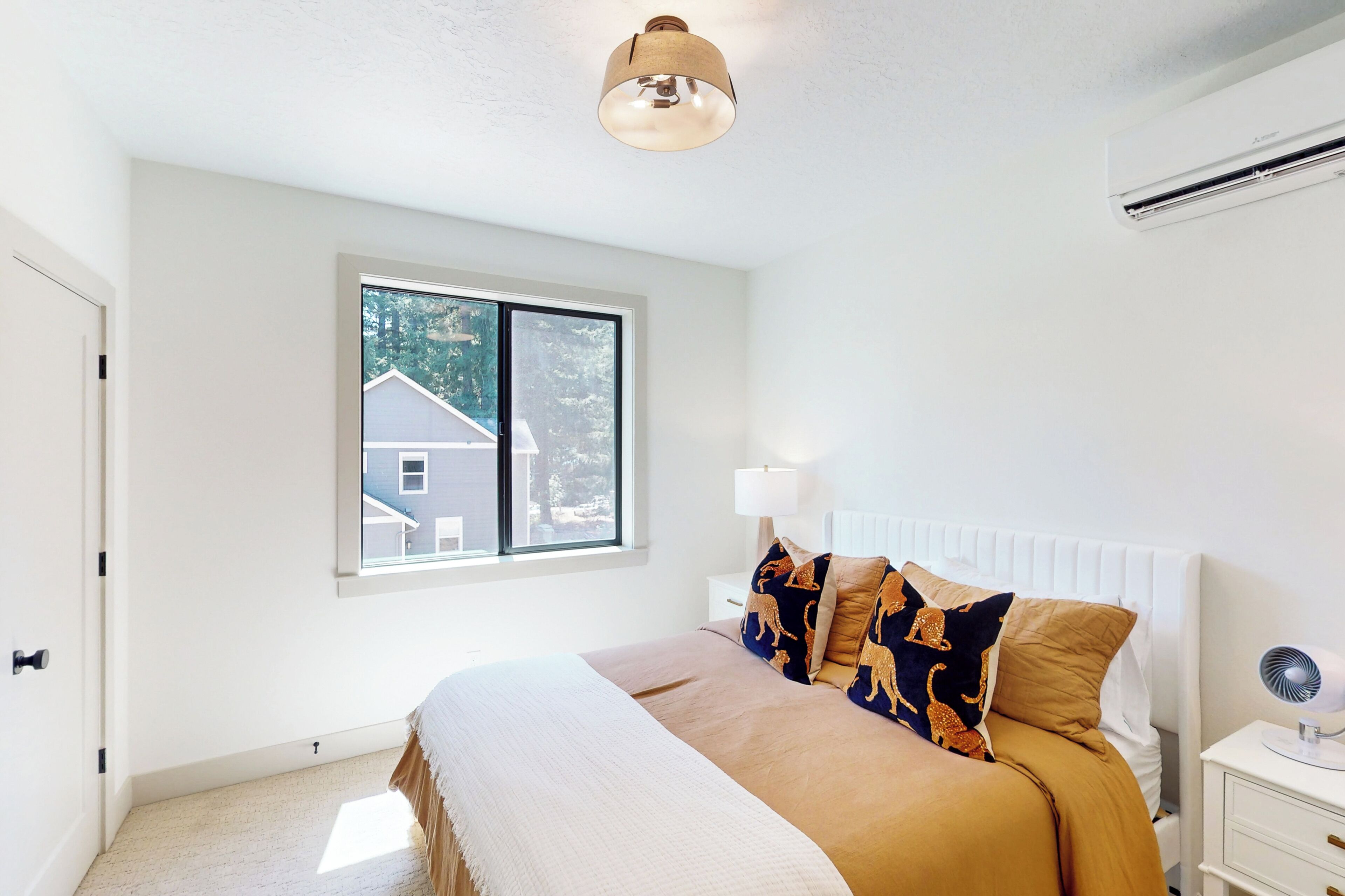 A bedroom with a large bed, a window with a view of a house, a bedside table with a lamp, and an air conditioning unit on the wall.