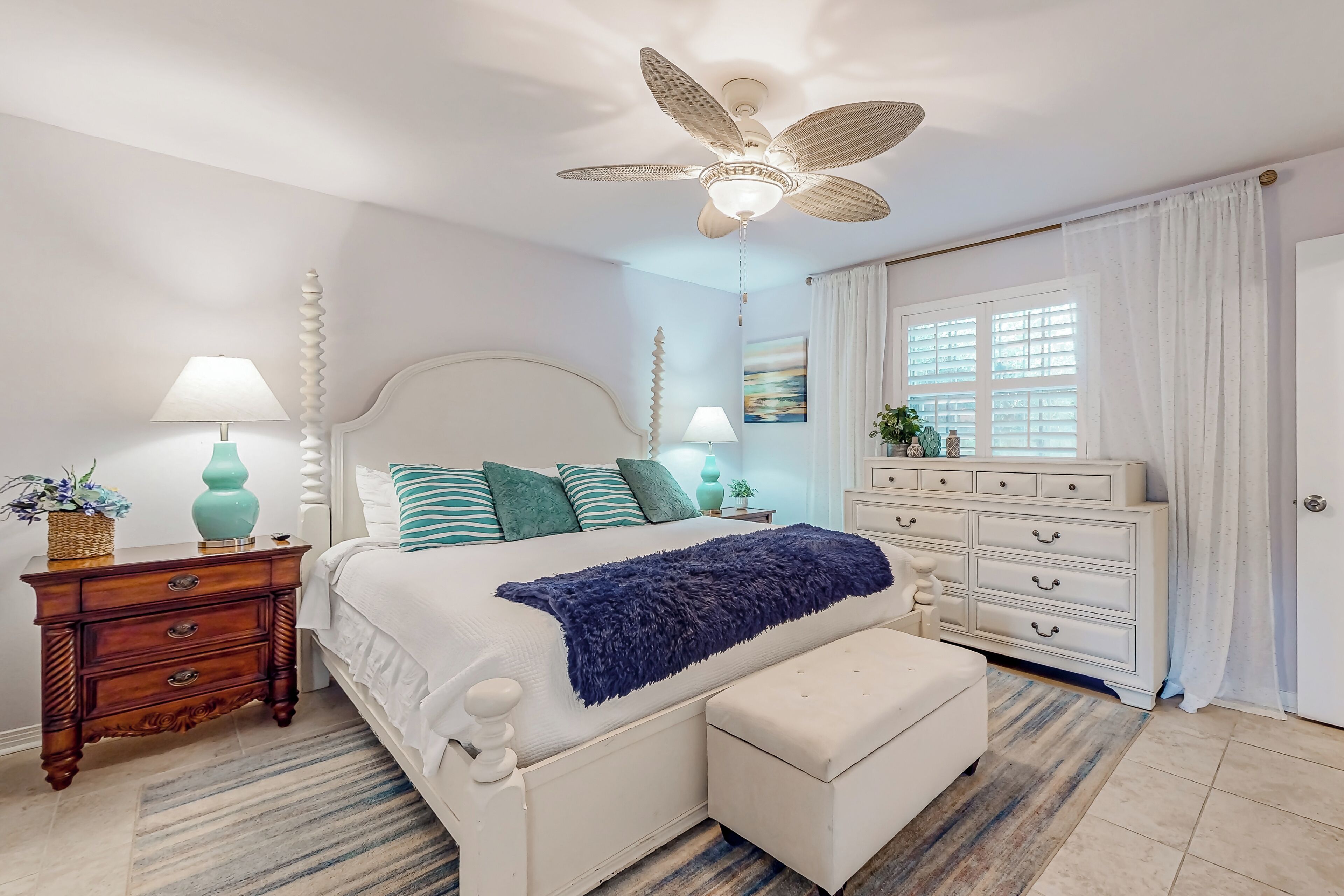 Un dormitorio con cama, mesitas de noche, cómoda, ventilador de techo y ventana con cortinas.