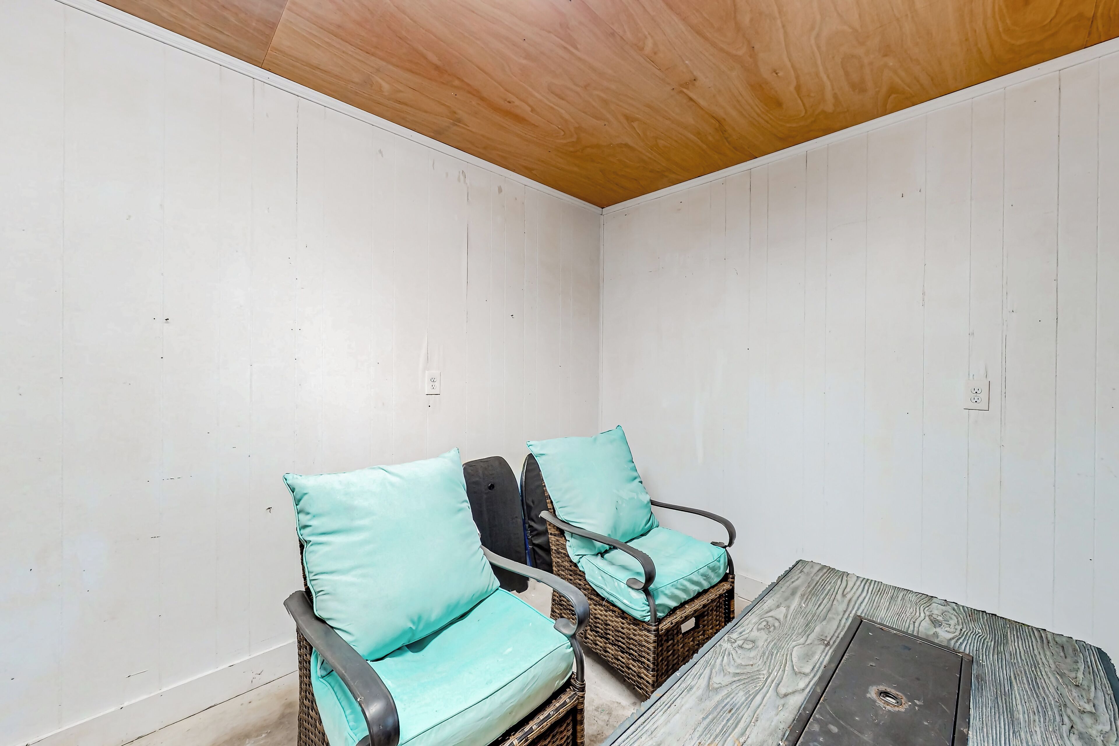 A corner room with two wicker chairs with teal cushions, a small table, and a wooden ceiling.