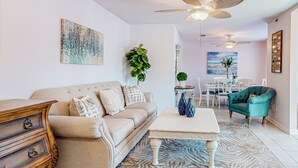 A living room with a beige sofa, a wooden dresser, a white coffee table, and a teal armchair. There are ceiling fans, a dining table with chairs, and a large window with a view.