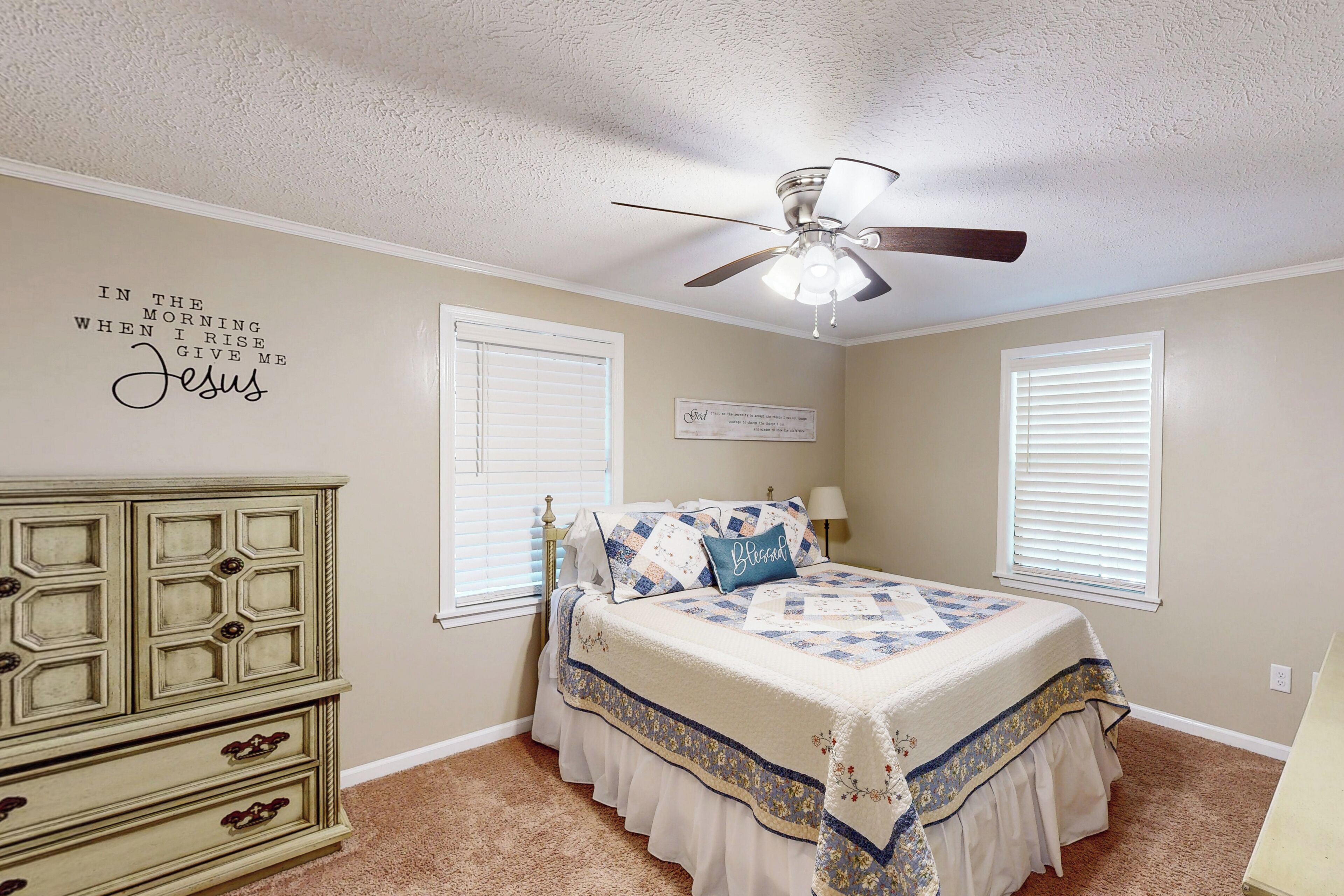 A bedroom with a bed, dresser, ceiling fan, and a wall quote.