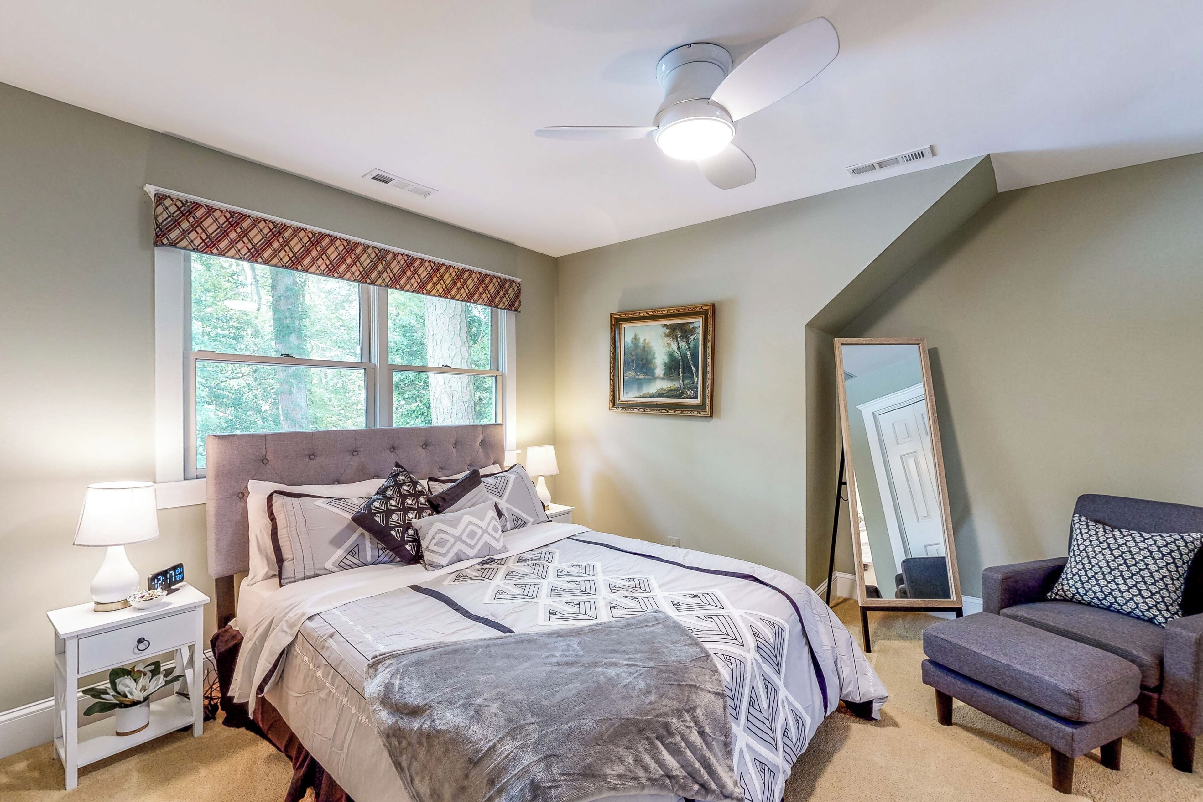 A bedroom with a bed, bedside tables, a chair, a ceiling fan, a window, and a mirror.