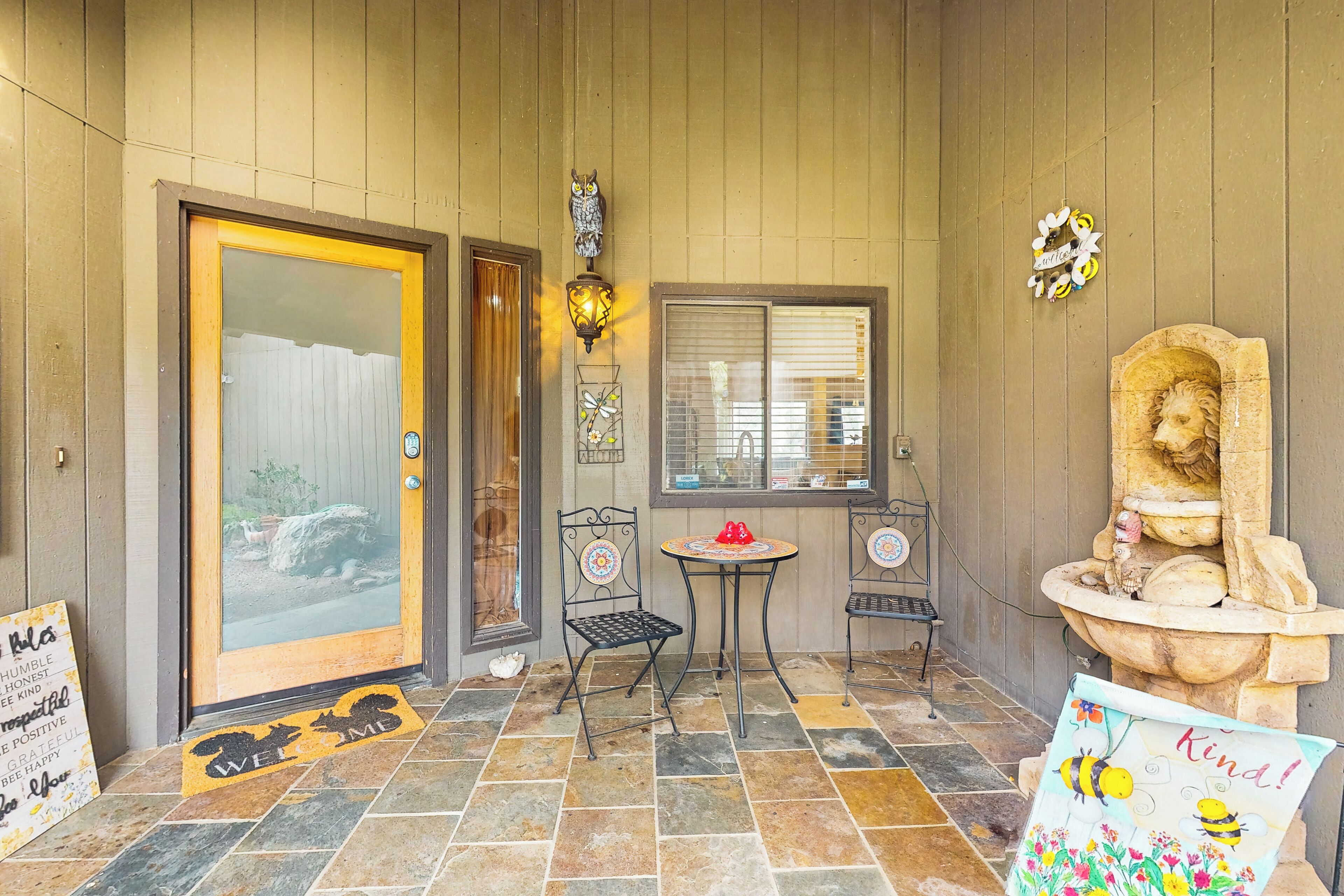 Une façade en bois avec une porte vitrée, une petite table avec des chaises, un lampadaire décoratif en forme de hibou et un paillasson orné d’un motif représentant un chien et un chat.