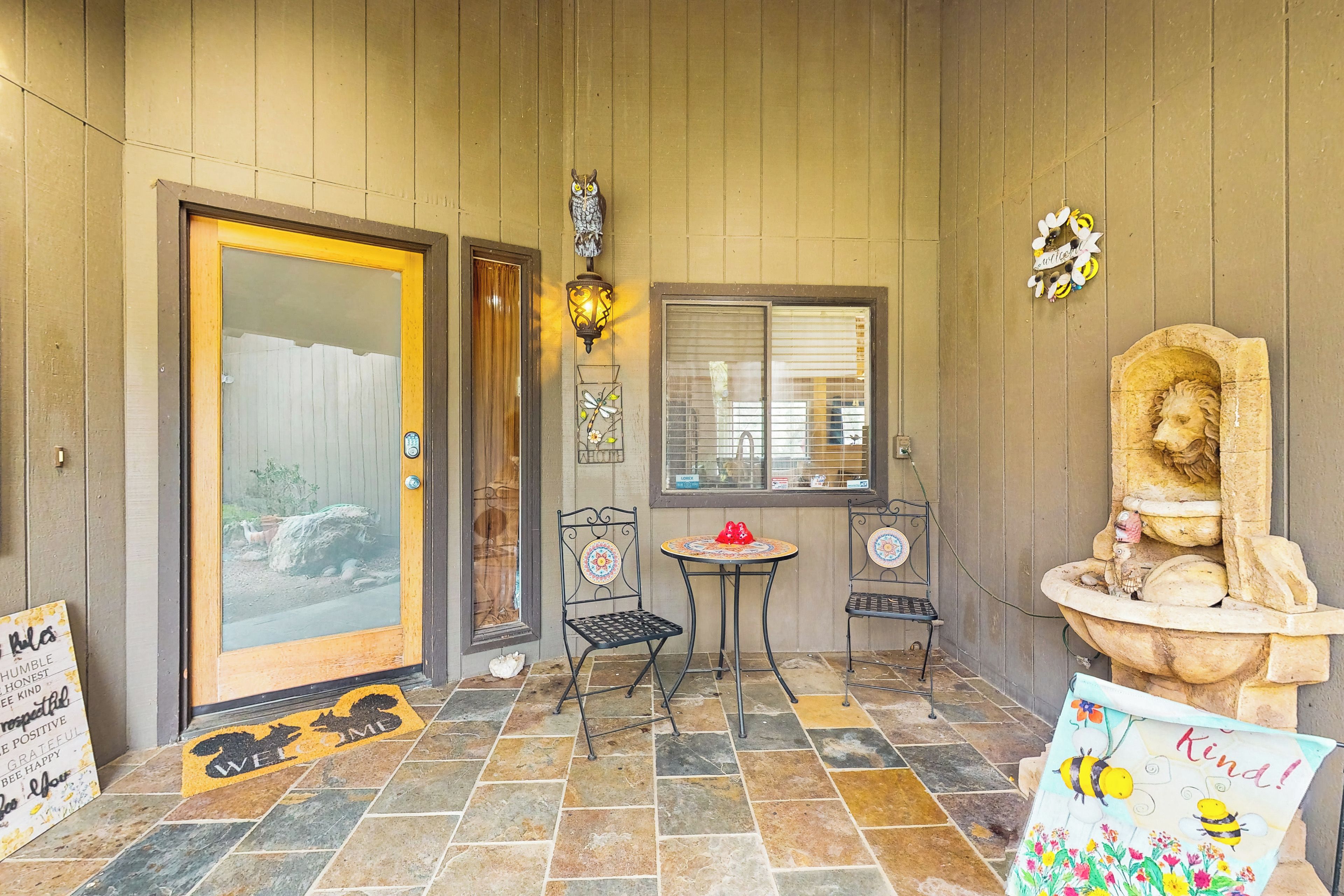 A wooden exterior with a glass door, a small table with chairs, a decorative owl light, and a welcome mat with a dog and cat design.