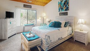 Una camera da letto con soffitto in legno, un letto con lenzuola blu e bianche, una televisione su un comò e una finestra vista alberi.