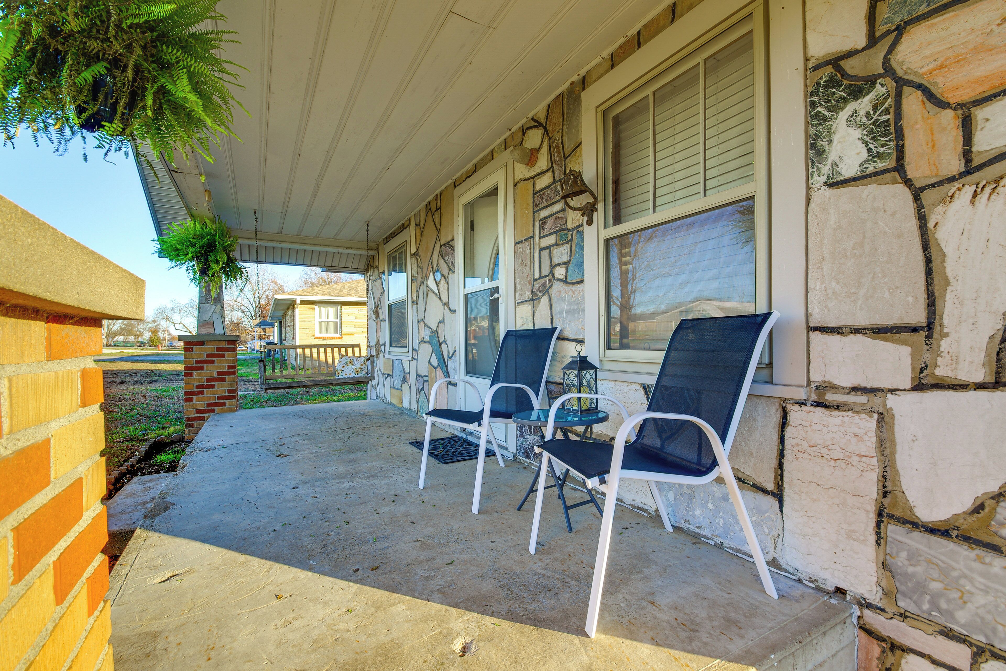 A covered porch with two chairs and a table, stone walls, and a hanging plant.