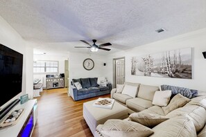 A spacious living room with a large flat-screen TV, a ceiling fan, and a dining area with a kitchen in the background.