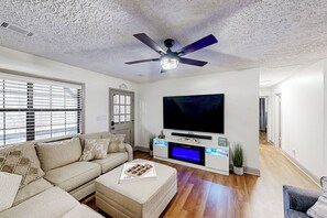 A living room with a sectional sofa, a flat-screen TV, a ceiling fan, and a wooden floor.