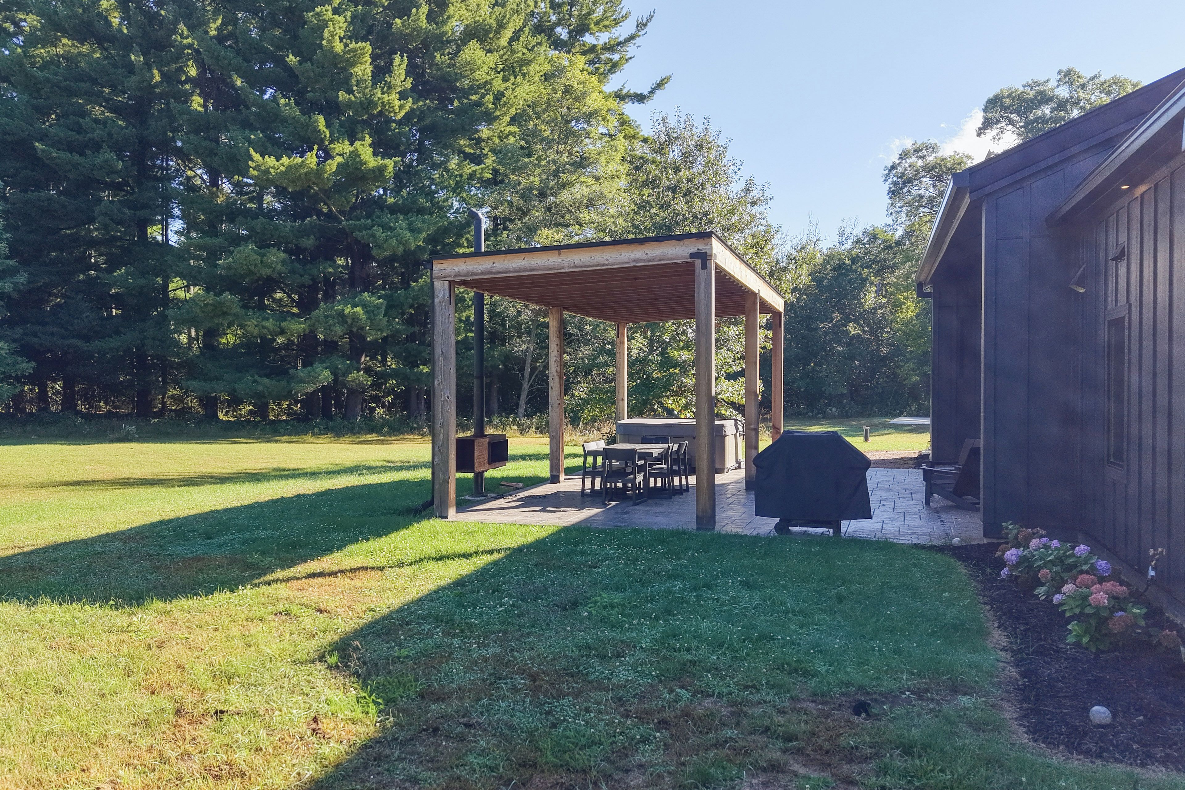 A covered outdoor seating area with tables and chairs, a barbecue grill, and a wooden building in the background.