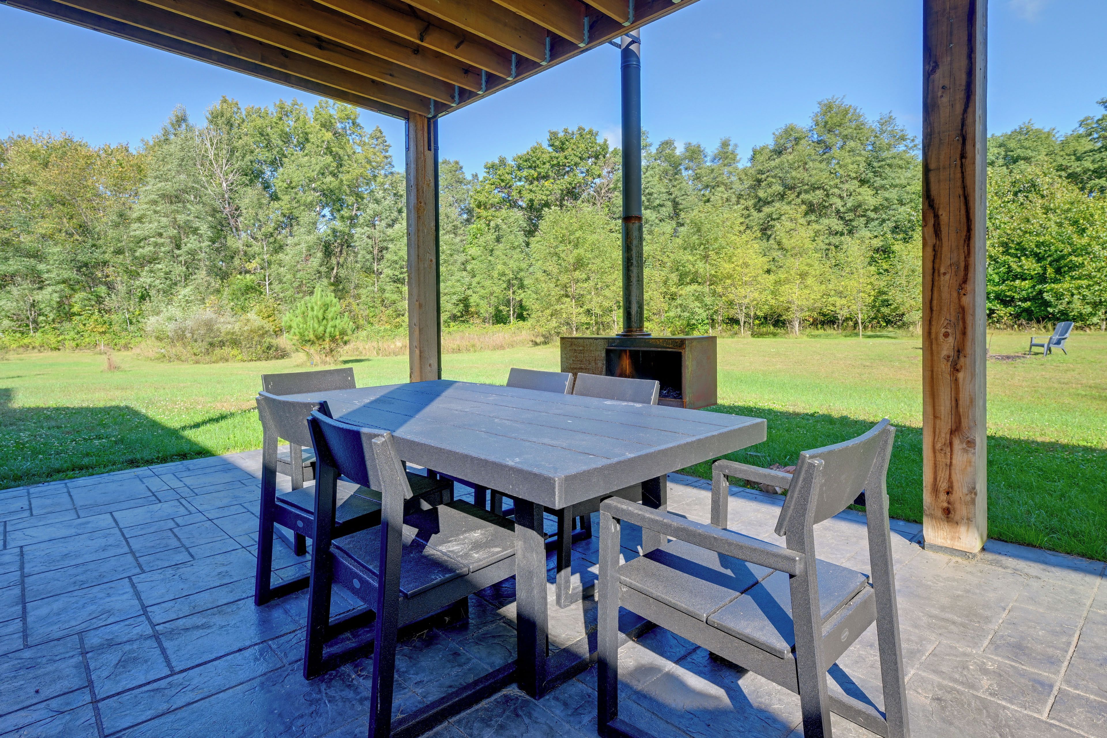 An outdoor patio with a wooden table and chairs, a chimney, and a grassy area with trees in the background.