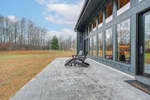 A modern building with large windows, a paved patio, and a chair outside. - Modern Necedah Home: Hot Tub, Woods & Lake Access (Necedah)