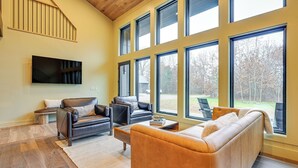 Ein geräumiges Wohnzimmer mit großem Fenster, Flachbildfernseher, Ledersofas und Holzboden.