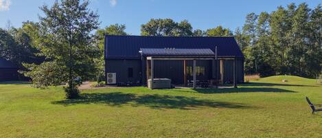 A single-story building with a covered patio area, surrounded by a grassy field and trees.