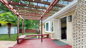 Eine überdachte Terrasse mit Holz-Pergola und Lichterkette, eine Palme und ein Ziegelhaus mit geöffneter Tür.