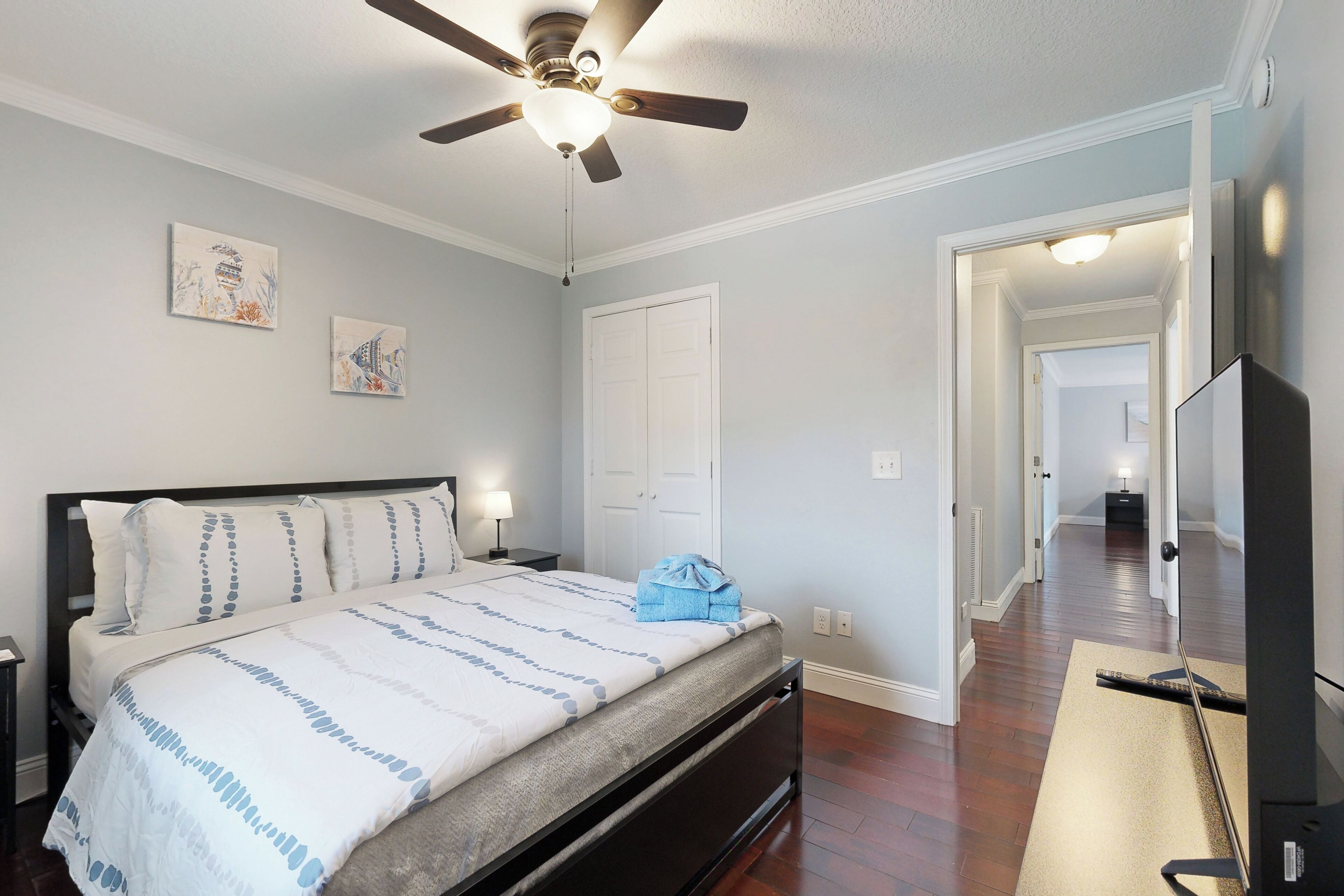 Un dormitorio con una cama, ventilador de techo, una obra de arte en la pared y un televisor sobre un mueble.