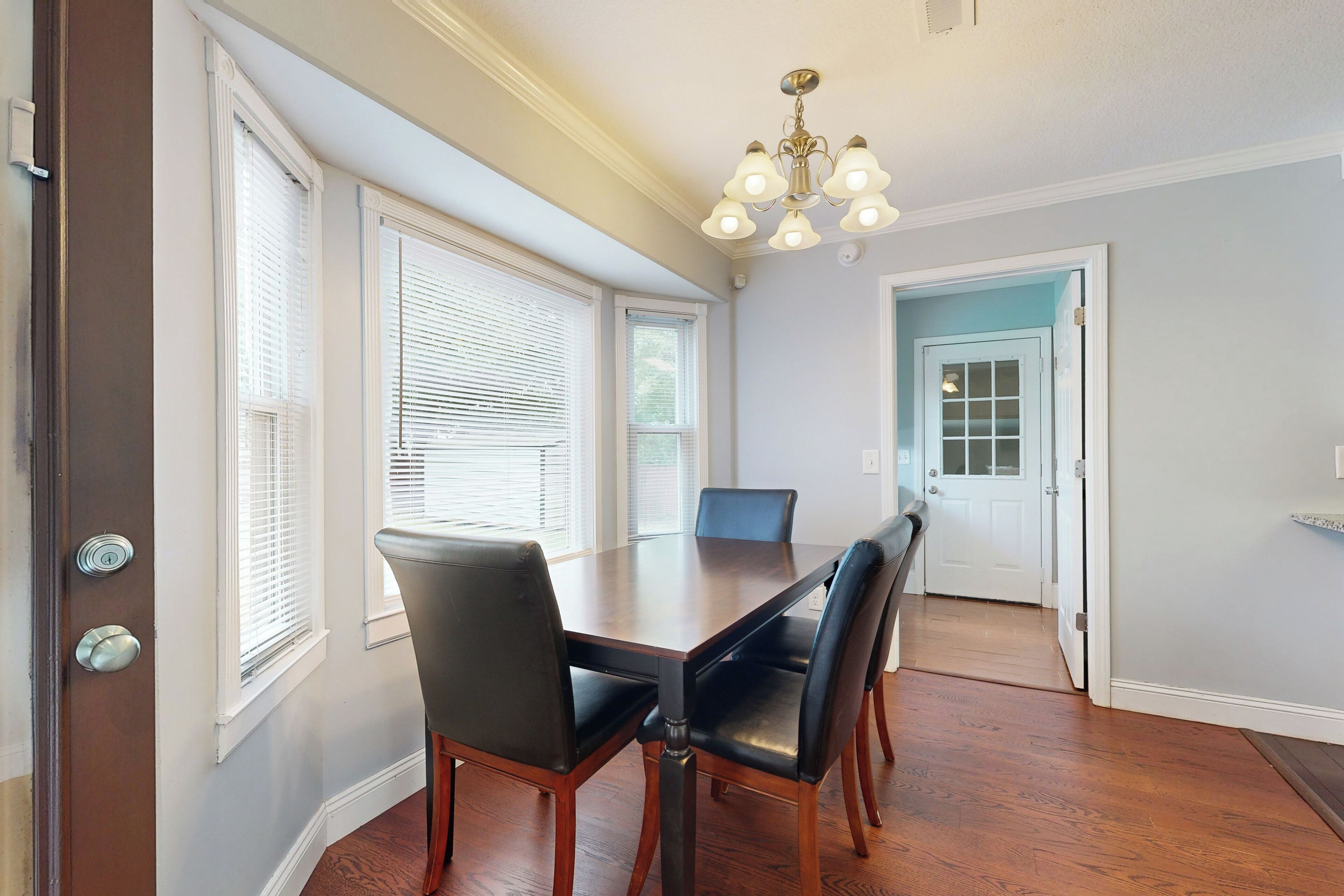 A dining area with a wooden table and chairs, large windows with blinds, and a chandelier.