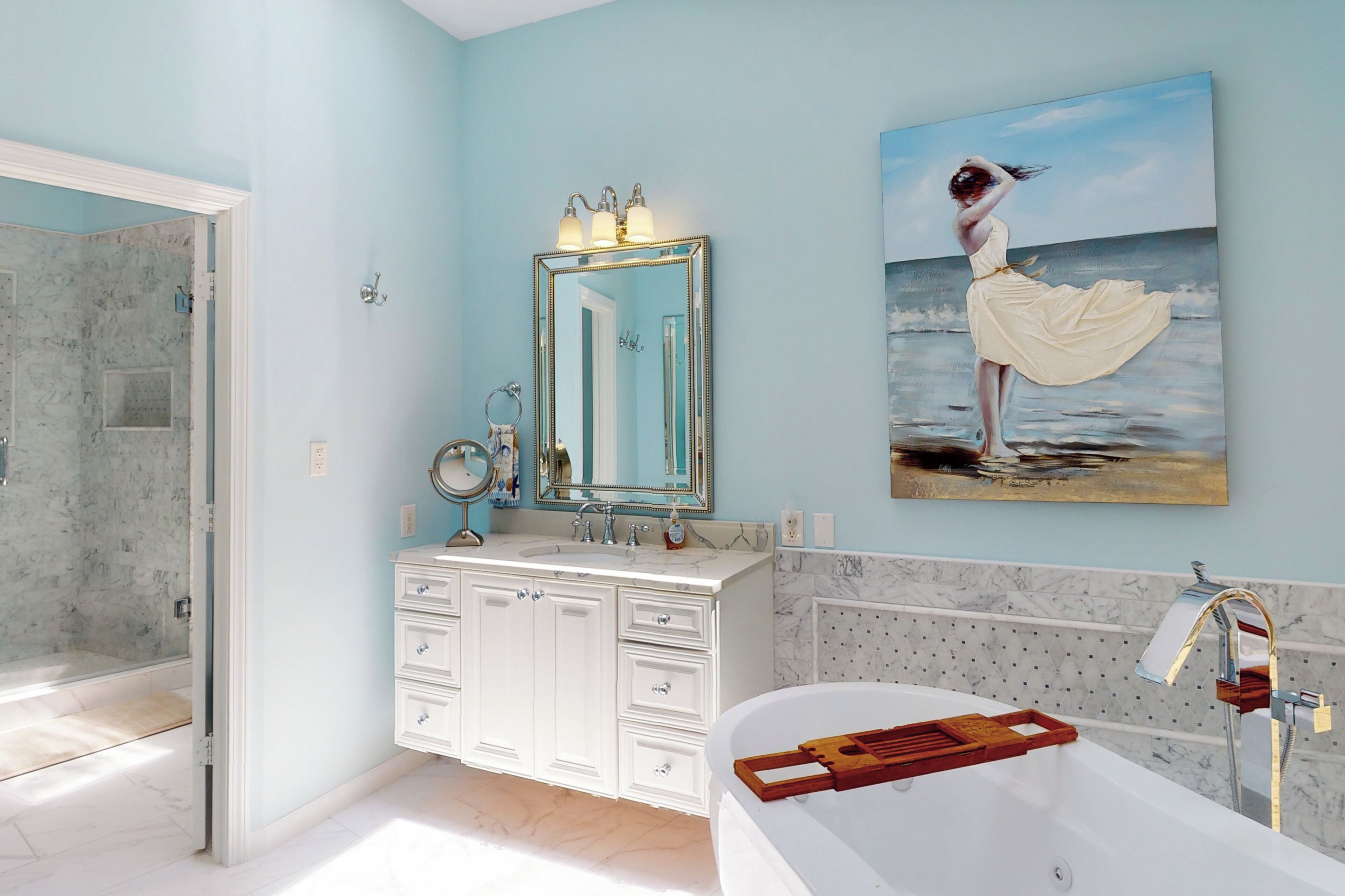 Un baño con un espejo grande, una bañera y un cuadro de una mujer junto al mar.