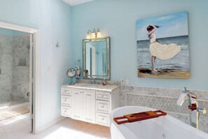 Un baño con un espejo grande, una bañera y un cuadro de una mujer junto al mar.